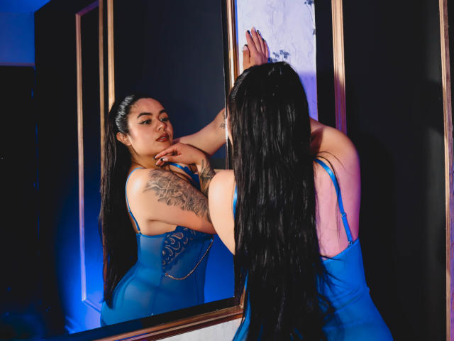 EveWillis with long black hair, blue dress, tattooed arm, touching mirror, reflecting her face and tits, standing against dark wall, blue lighting, looking serious, hands up.