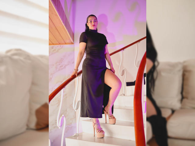 ElizabethDupont with black dress, high heel platform shoes, and one leg raised on white stairs, showing her thigh. She's standing by a red railing, with purple lighting.