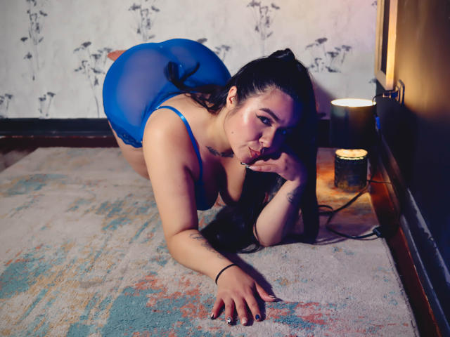 EveWillis on all fours in a blue sheer lingerie set, showing her ass and tits, with long black hair, a tattoo on her arm, and a candle beside her.