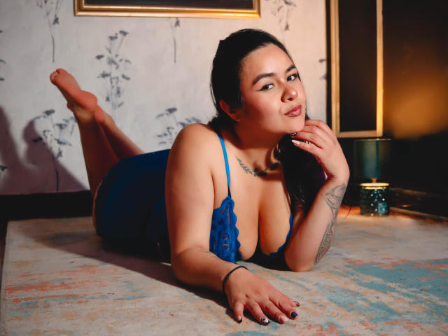 EveWillis, laying on a rug, wearing a blue lace lingerie set, showing her tits, with one hand near her face, legs up, tattoo on arm, dark hair, light skin.
