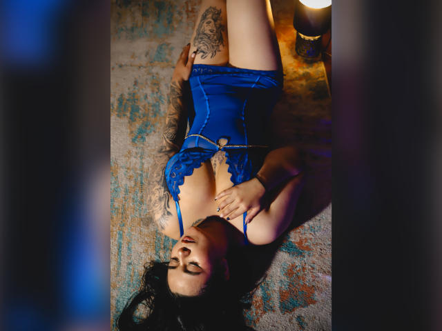 EveWillis, with tattoos on her arms, lies on a rug, wearing a blue lace lingerie top, her hands on her tits, eyes closed, legs up, and a lamp nearby.