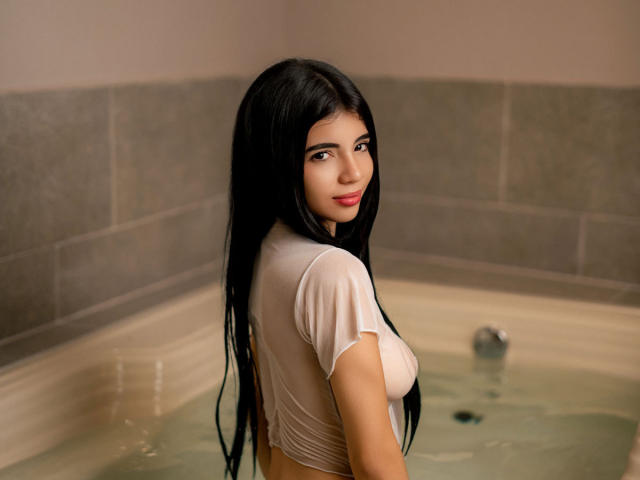 RoxanneeSecret with long black hair, wet white t-shirt clinging to her boobs, standing in a bathtub, looking over her shoulder with a slight smile, in a bathroom.