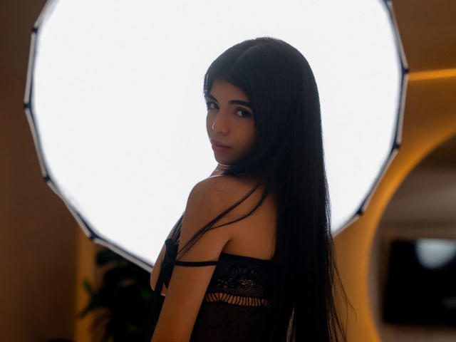 RoxanneeSecret with long black hair, looking back over her shoulder, wearing a black lace top, showing her tits, standing in front of a bright circular light.
