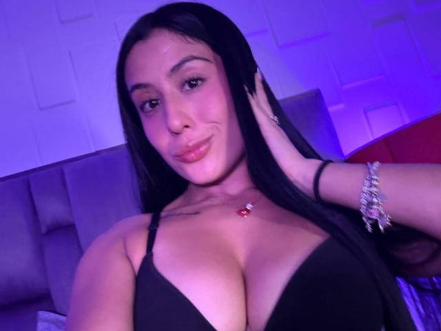 CassieFox with long black hair, smiling, wearing a black bra that shows her big tits, a red necklace, and a silver bracelet, against a purple-lit background.