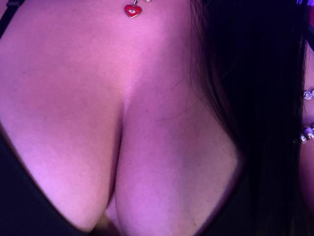CassieFox's big, bare boobs with a small red heart necklace between them, her black hair partially covering her right side, under purple lighting.