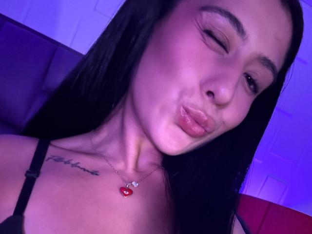 CassieFox with black hair, puckering her lips, wearing a black strap top, red heart necklace, and tattoo on her right chest, under purple lighting.