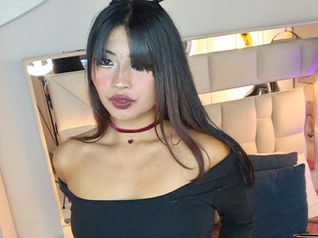 IssaJones with long black hair, bangs, off-shoulder black top, red choker, pink lipstick, medium tits, light skin, in a bedroom with a white, tufted headboard.