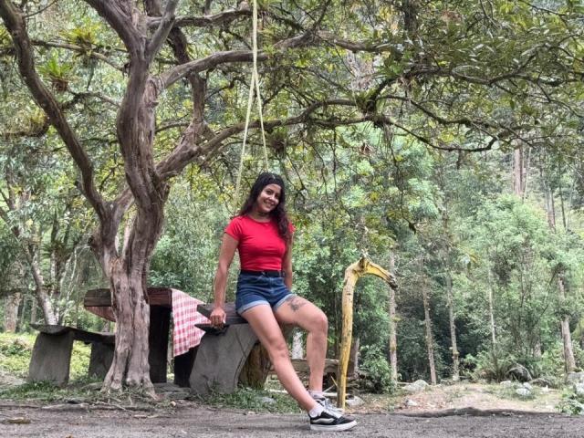 ElectraRoss with long dark hair, red shirt, denim shorts, and black sneakers, sitting on a tree swing, smiling, forest background, yellow excavator on right, tree on left.