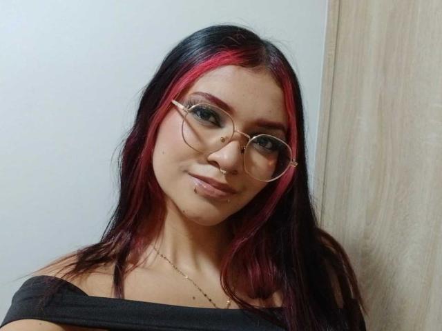 AriaRosel with black and red hair, glasses, nose ring, and off-shoulder black top, showing a subtle smile. Her tits are small, and she has a cute face.