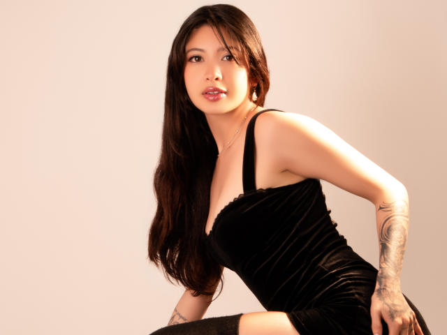 Megan, con el pelo largo y negro, vestida con un vestido de terciopelo negro, tatuaje en el brazo derecho, inclinada hacia adelante, mostrando pequeños senos, sonrisa suave, lápiz labial rosa, piel clara, pose sexy..
