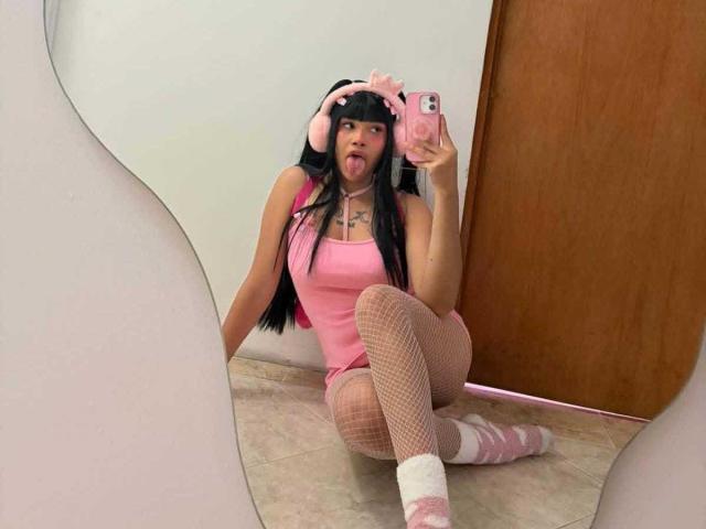 LiMiuu with long black hair, pink tank top, fishnet stockings, pink socks, and pink headphones, sticking her tongue out, kneeling in front of a mirror, holding a pink phone.