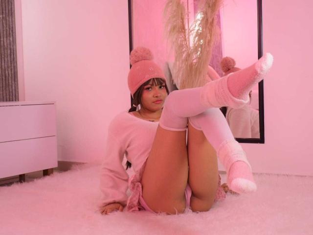 LiMiuu is sitting on pink fluffy carpet, wearing a pink knit hat, pink sweater, and white knee socks, with her legs up, showing her pussy, in a pink-lit room.