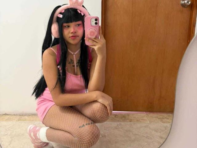 LiMiuu in pink outfit, fishnet stockings, pink headphones, squatting, mirror selfie, pink phone, white wall, wooden door, tattoo on left thigh, long black hair, pink sneakers.
