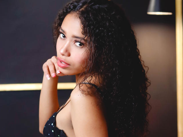 ViolettExposito with curly black hair, light brown skin, and pink lips, looking over her shoulder with a slight pout, hand near her mouth, wearing a black, polka-dotted top.