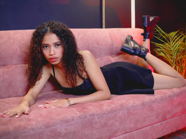 ViolettExposito with curly brown hair, lying on pink couch in black dress, showing her ass, wearing black high heels, and a gold bracelet, looking at camera.