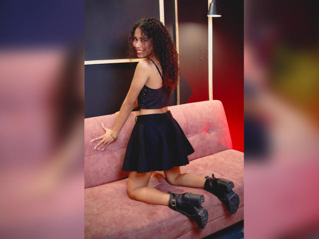 ViolettExposito kneels on a pink sofa, wearing a black crop top, black skirt, and black boots, with curly hair and a smile, showing her ass.