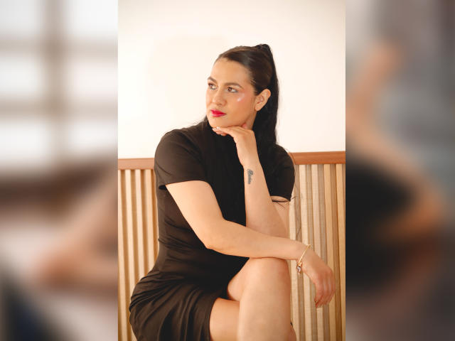 Elizabeth Dupont, con rossetto rosso, vestito nero e coda di cavallo, seduta con il mento appoggiato sulla mano, che mostra un piccolo tatuaggio sul polso, e il sedere leggermente visibile..