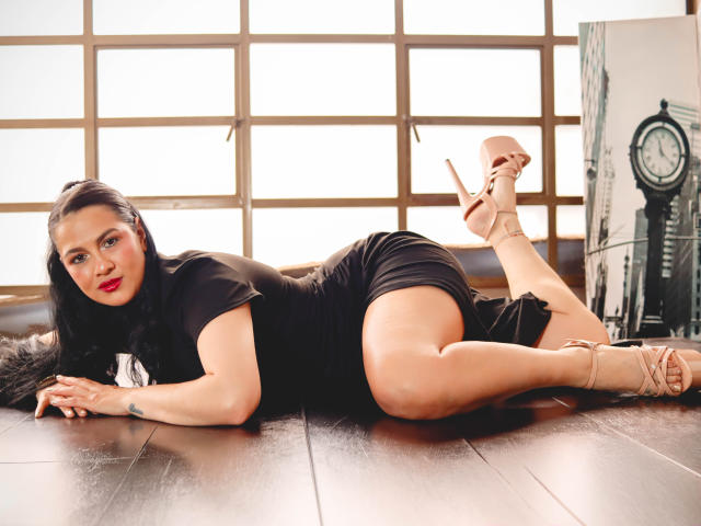 ElizabethDupont is laying on her side with her ass up, wearing a black dress and tan high heels, her red lips contrasting with her dark hair and fair skin.