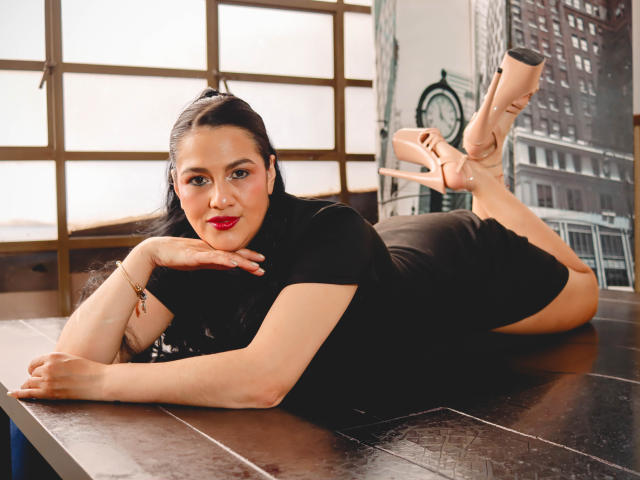 ElizabethDupont, lying on her stomach on a wooden table, wearing a black dress, pink heels, red lipstick, and a bracelet, with her hand under her chin, looking at the camera.