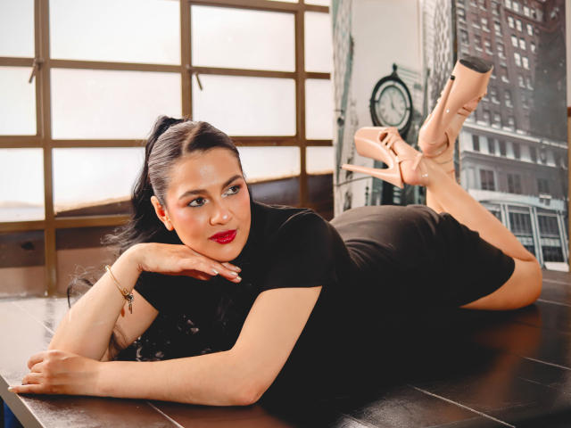 ElizabethDupont laying on her stomach, black dress, red lipstick, long black hair in a ponytail, beige high heels, butt up, legs crossed, hand under chin, wooden table.