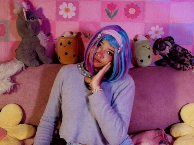 LiMiuu with blue and pink wig, purple and white hair, wearing a gray sweater, hand on cheek, surrounded by stuffed animals, pink wall, purple and pink sofa.