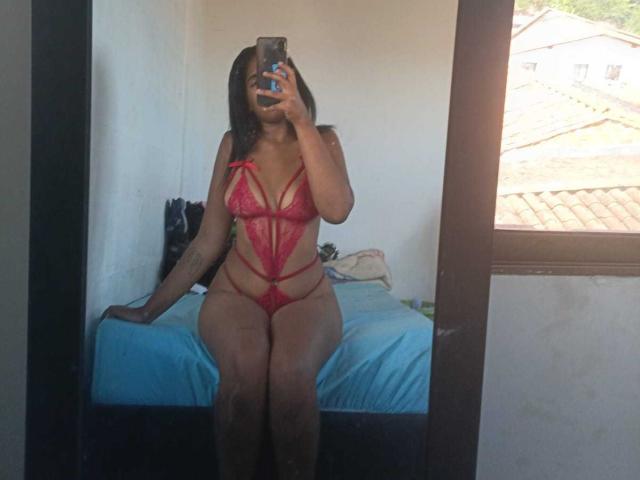 MiaStClaire wearing a red lace lingerie set with strappy details, sitting on a bed, taking a mirror selfie with her phone, showing her dark skin and black hair.