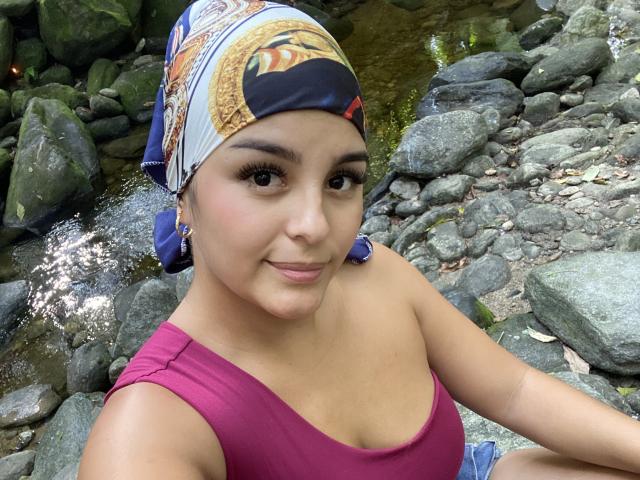 TalianaSquirt with tan skin, brown eyes, and long lashes, wearing a colorful headscarf and pink top, sitting by a rocky stream. Her tits are small and she's smiling.