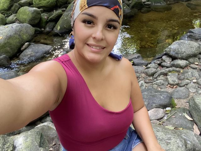 TalianaSquirt, with medium tits in a pink tank top, smiling at the camera, wearing a colorful headscarf, sitting on rocky ground by a clear stream.