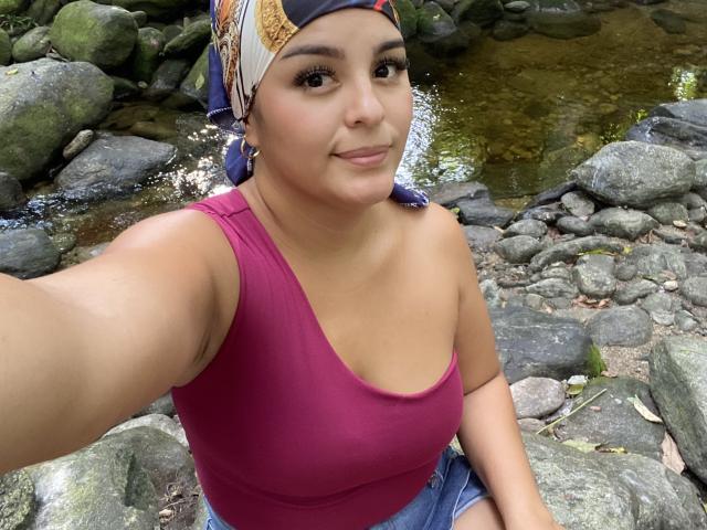 TalianaSquirt, met een gemiddelde borstmaat en een roze tanktop, zit op rotsen bij een beek, draagt een kleurrijke hoofddoek, glimlacht en maakt een selfie..