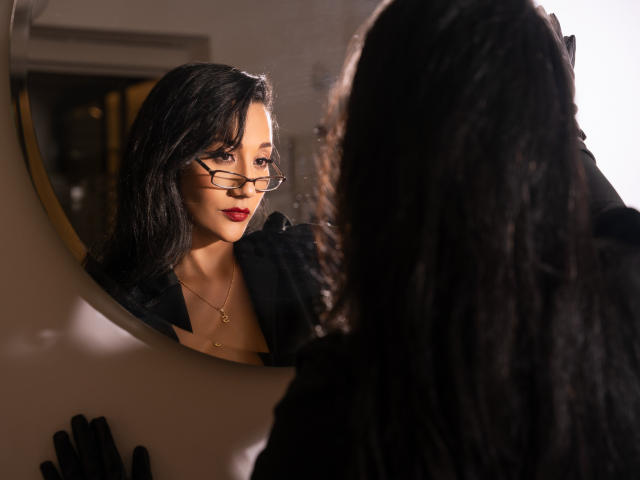 Kourtney Parker mit Brille, schwarzem Haar, rotem Lippenstift und schwarzem Kleid, die in einen Spiegel schaut. Im Spiegelbild sind ihr Gesicht und ihre Brust zu sehen. Sie trägt eine kleine goldene Halskette, und im Vordergrund sind schwarze Handschuhe zu sehen..