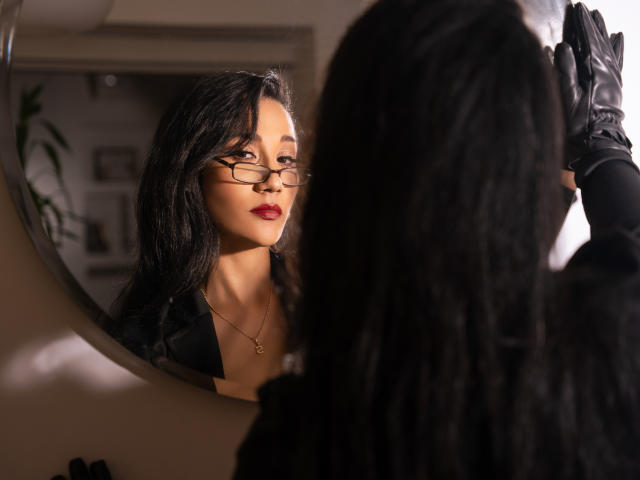 KourtneyParker with glasses, black gloves, and a necklace, looking in a mirror with her ass slightly visible in the reflection, has long black hair and red lipstick.