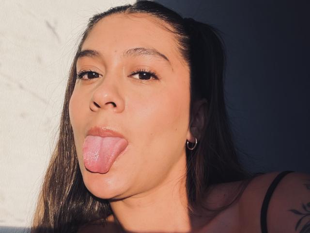 ThaliaAres with her tongue out, looking up, has brown hair in pigtails, small hoop earrings, and a tattoo on her right shoulder, in a sunny, well-lit portrait.