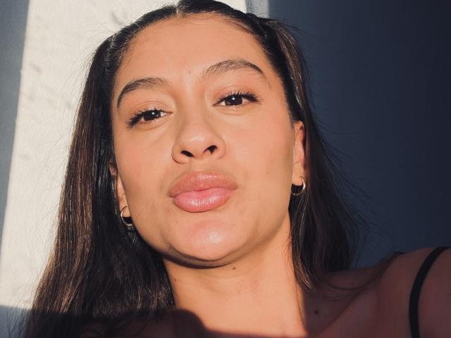 ThaliaAres with pouty lips, brown eyes, and dark straight hair, wearing hoop earrings, sunlight highlighting her tan skin and subtle makeup. Boobs partially visible in black top.