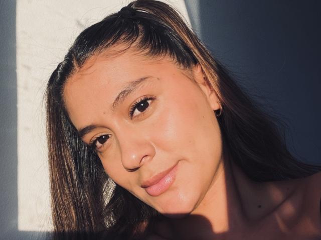 ThaliaAres has brown eyes, long dark hair in pigtails, light brown skin, small nose, subtle smile, and small black earrings, with sunlight highlighting her face.