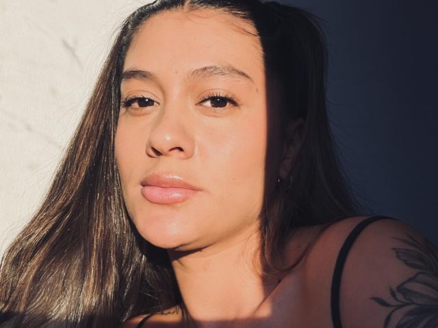 ThaliaAres has long brown hair, light brown eyes, and pink lips with a subtle tattoo on her right shoulder. She's wearing a black spaghetti strap top.