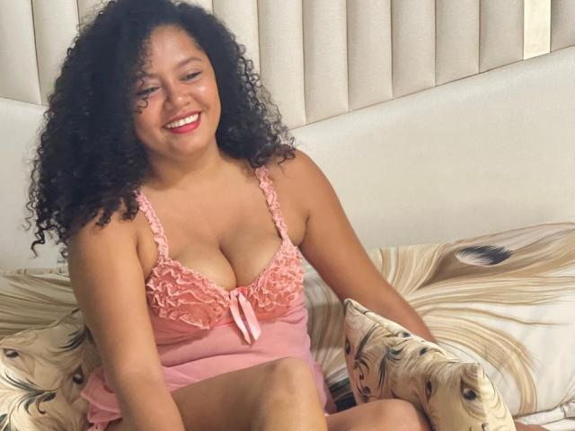 Curvy DaisyCrespa with curly black hair, smiling, wearing pink lace lingerie that shows her tits, sitting on a bed with floral pillows, her pink nipples slightly visible.
