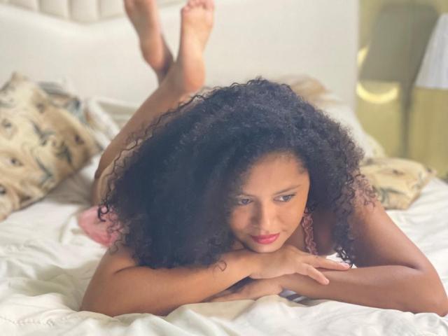DaisyCrespa with curly black hair, lying on a bed, legs up, arms folded, looking at camera, wearing pink lingerie, feet in air, white sheets, soft light.