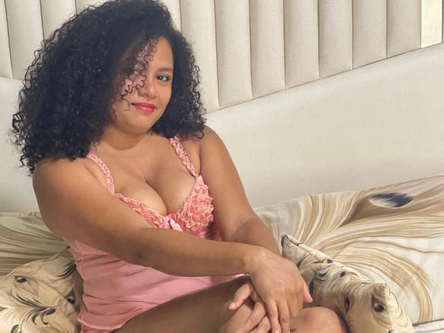 DaisyCrespa with curly black hair, pink ruffled lingerie, and red lipstick, sitting on a bed with beige patterned sheets, showing her tits and smiling.
