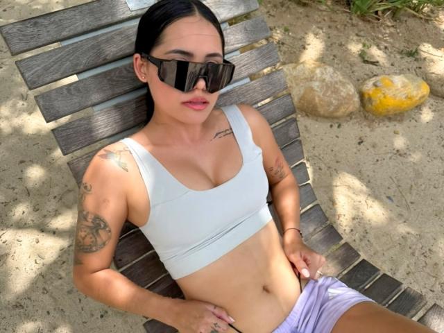 KatyJhoans with black hair, wearing sunglasses, a white sports bra, and purple shorts, lying on a wooden lounge chair, showing tattoos and a flat stomach.