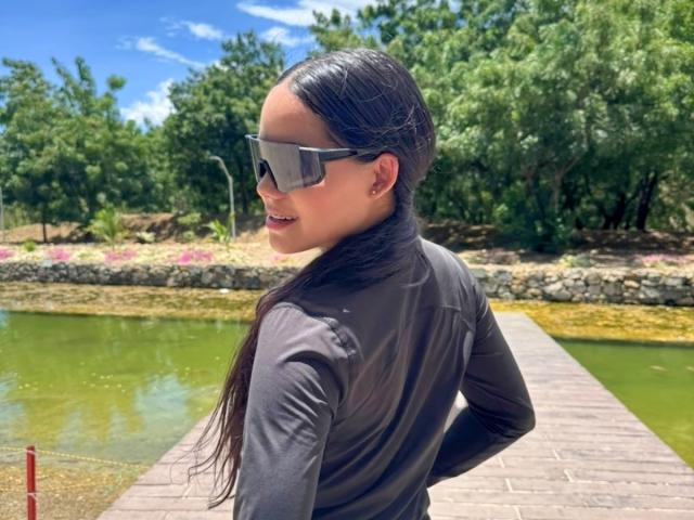 KatyJhoans with long dark hair, wearing sunglasses, a gray long-sleeve shirt, and small earrings, standing on a wooden bridge with a green pond in the background.