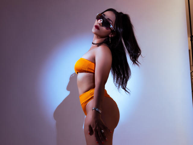 KourtneyParker with long black hair, wearing orange strapless bikini, standing sideways, showing her ass, shadow on wall, blue light, glasses, choker, bracelet, looking up, sexy pose.