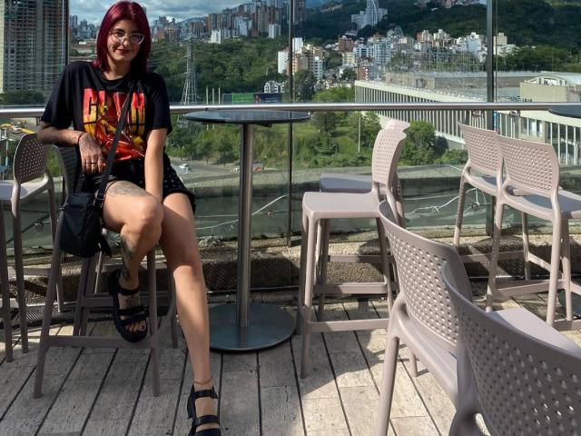 NyxVale with red hair, glasses, black t-shirt, shorts, and black sandals, sitting on a chair with legs crossed, showing a tattoo on her right thigh.
