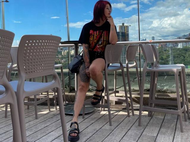 NyxVale with red hair, black "Iron Maiden" shirt, black shorts, tattoos, sitting on chair, black sandals, holding bag, outdoor deck, clear sky, city skyline in background.