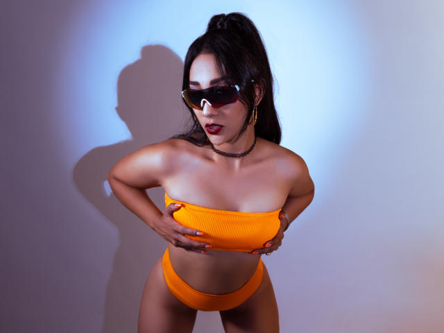 Kourtney Parker in een oranje bikini, die haar borsten vasthoudt, draagt rode zonnebril, een zwarte choker en een paardenstaart, met een schaduw op een witte muur, kijkt naar beneden, heeft rode lippenstift op, heeft een gemiddelde borstmaat en een slank postuur..