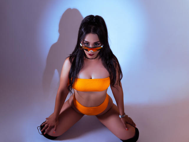 Kourtney Parker, com cabelo escuro, vestindo um top bandeau laranja e calcinha de cintura alta combinando, agachada, com as mãos nos joelhos, máscara laranja, sombra na parede, meia-calça preta alta, relógio de pulso..