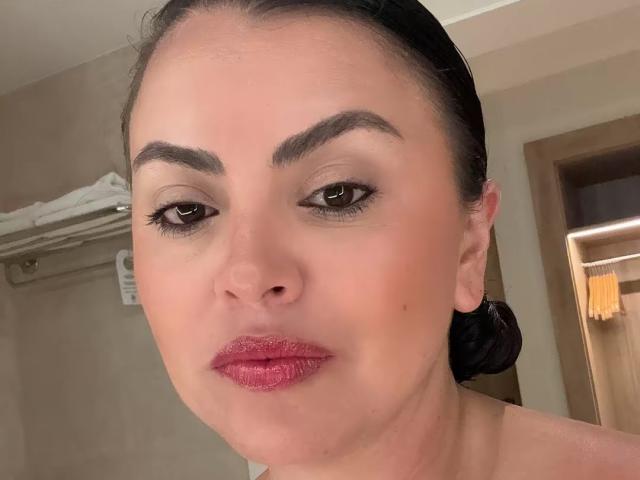 CharlotteDevereaux has dark brown eyes, thick black eyebrows, and pink lipstick on her slightly parted, glossy lips. Her black hair is pulled back, and she has a fair skin tone.