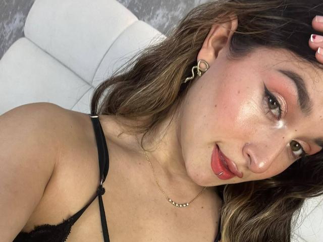 MilaGonz has brown eyes, pink eyeshadow, red lipstick, and a gold hoop earring. She's wearing a black lace bra, her tits visible, with her head tilted.