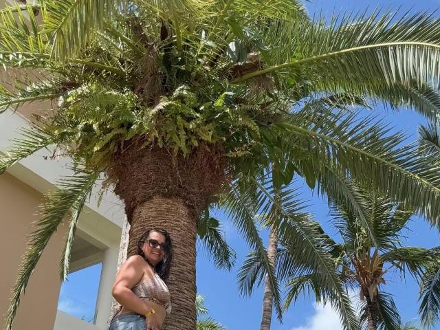 CharlotteDevereaux with dark hair and sunglasses, standing under a tall palm tree, wearing a beige lace top, blue jeans, and a yellow wristband, looking down.