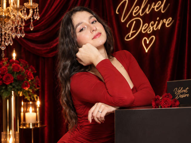 MilaGonz with long wavy brown hair, red dress, and ring, leaning on black box with red roses, looking cute and sexy, surrounded by candles and rose bouquet.