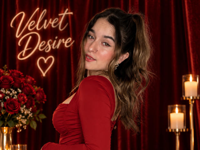 MilaGonz with wavy brown hair in a red dress, showing her tits, smiling at the camera, red roses, candles, and velvet curtains. She's looking over her shoulder.